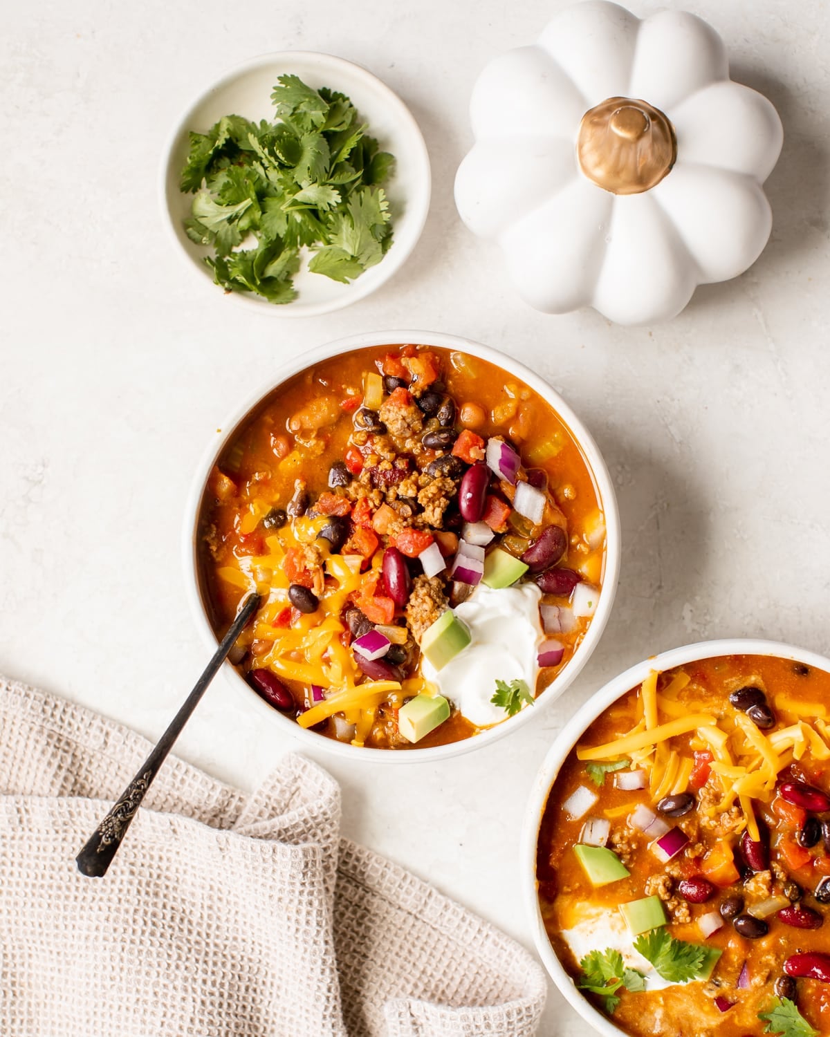 a white bowl filled with chili made with beans and garnished with red onion, cheese, cilantro, sour cream and avocado. 