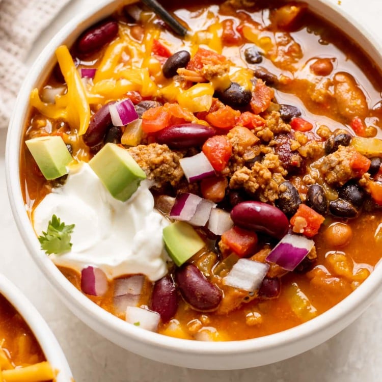 a white bowl filled with chili made with beans and garnished with red onion, cheese, sour cream and avocado.