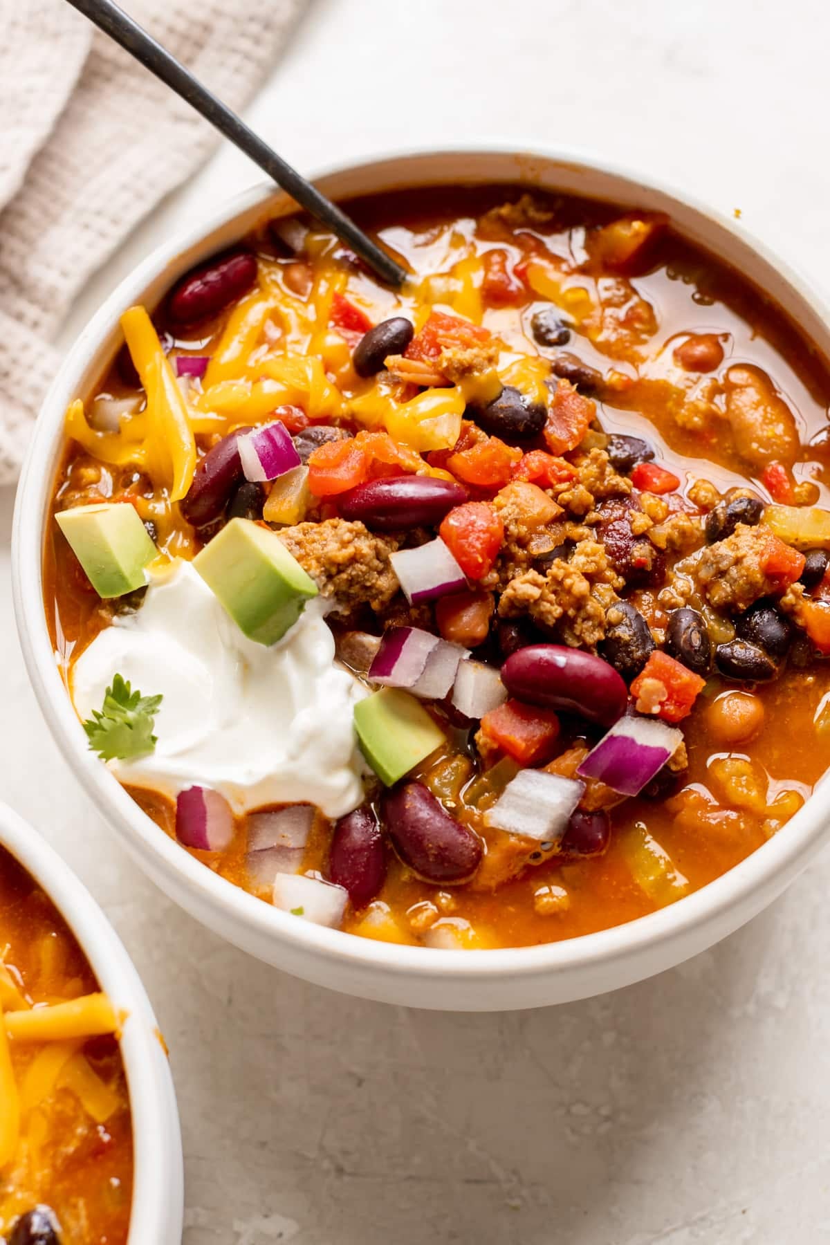 a white bowl filled with chili made with beans and garnished with red onion, cheese, sour cream and avocado.