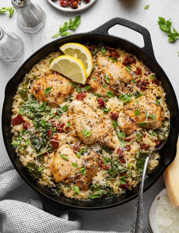 Overhead view of a cast iron skillet filled with creamy Tuscan chicken and rice, featuring golden-browned chicken thighs nestled in creamy Parmesan rice with spinach and chopped sun-dried tomatoes. Garnished with grated Parmesan, fresh parsley, and lemon slices, with additional Parmesan, sun-dried tomatoes, and seasonings styled around the skillet.
