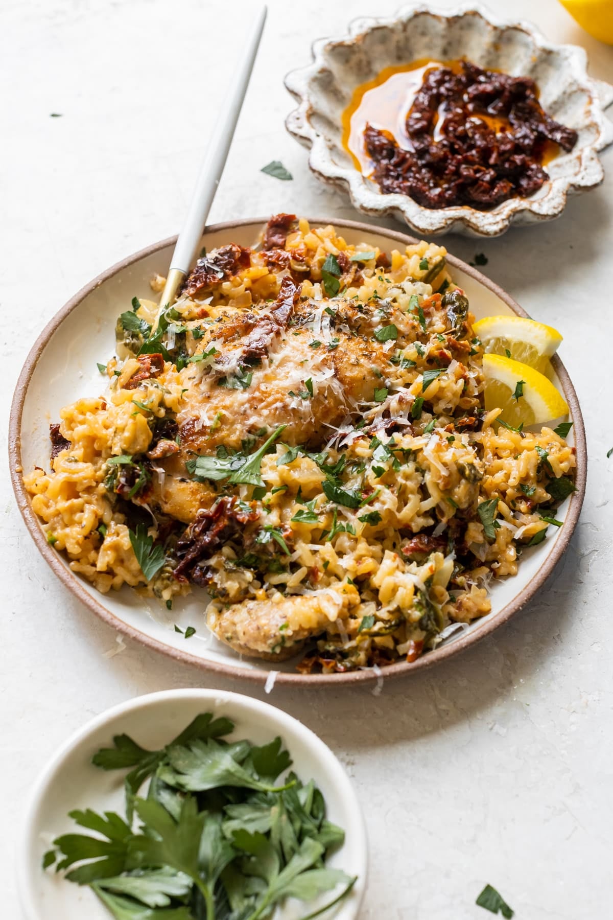 chicken and rice on a white plate garnished with parmesan cheese, sun dried tomatoes, parsley and lemon slices. 