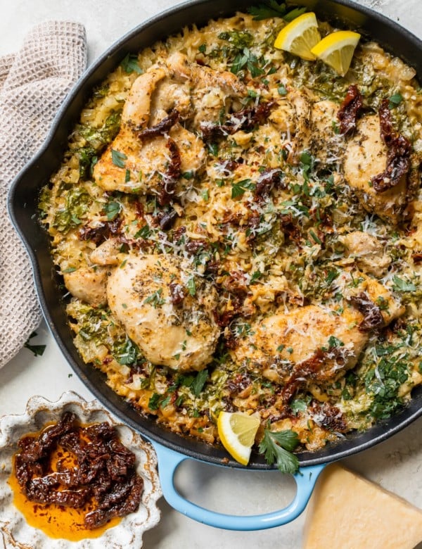 chicken and rice in a cast iron skillet garnished with parmesan cheese, sun dried tomatoes, parsley and lemon slices.