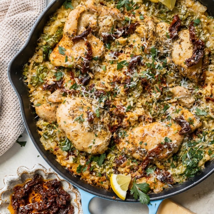 chicken and rice in a cast iron skillet garnished with parmesan cheese, sun dried tomatoes, parsley and lemon slices.