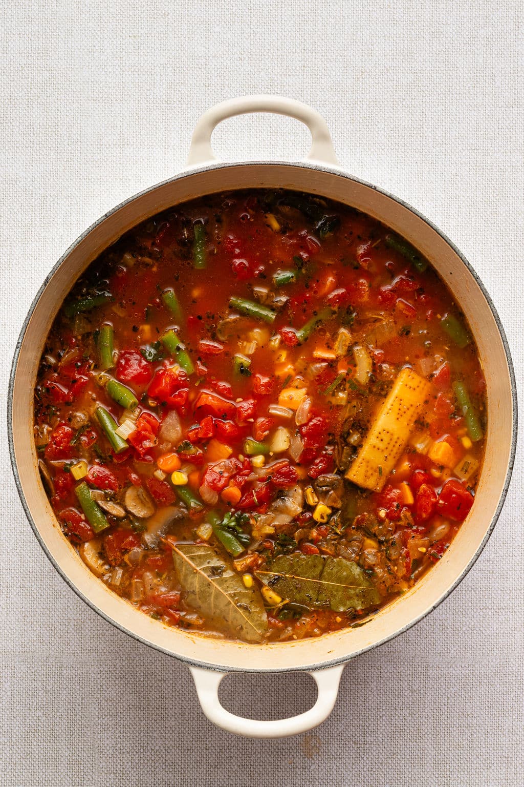 Overhead view of a Dutch oven filled with hearty vegetable soup, packed with tomatoes, carrots, green beans, corn, onions, and mushrooms, simmering with bay leaves and a parmesan rind in a rich broth.