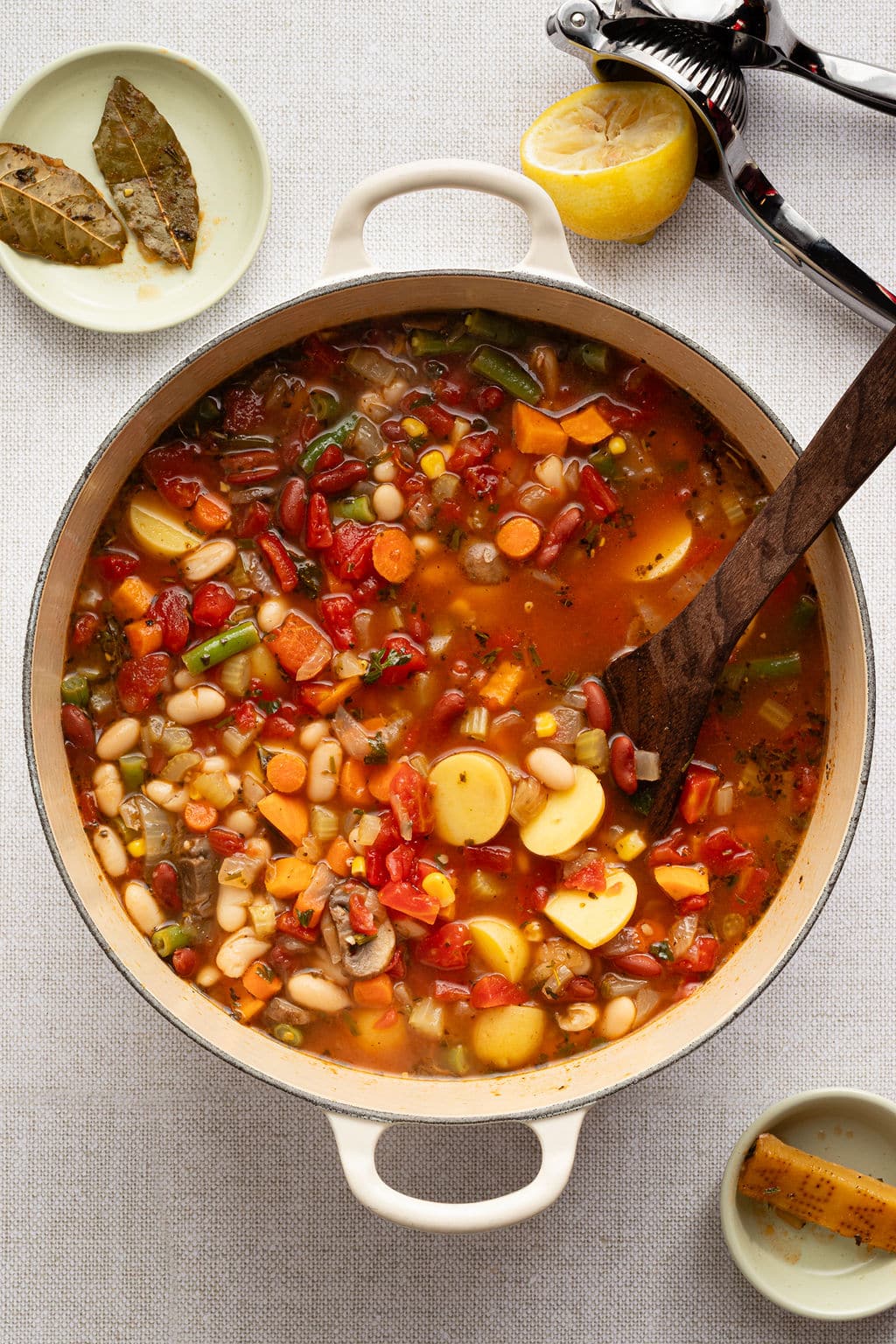 Large pot of hearty vegetable soup filled with colorful ingredients like carrots, potatoes, green beans, corn, tomatoes, and beans, simmering with bay leaves on the side and a lemon half nearby.