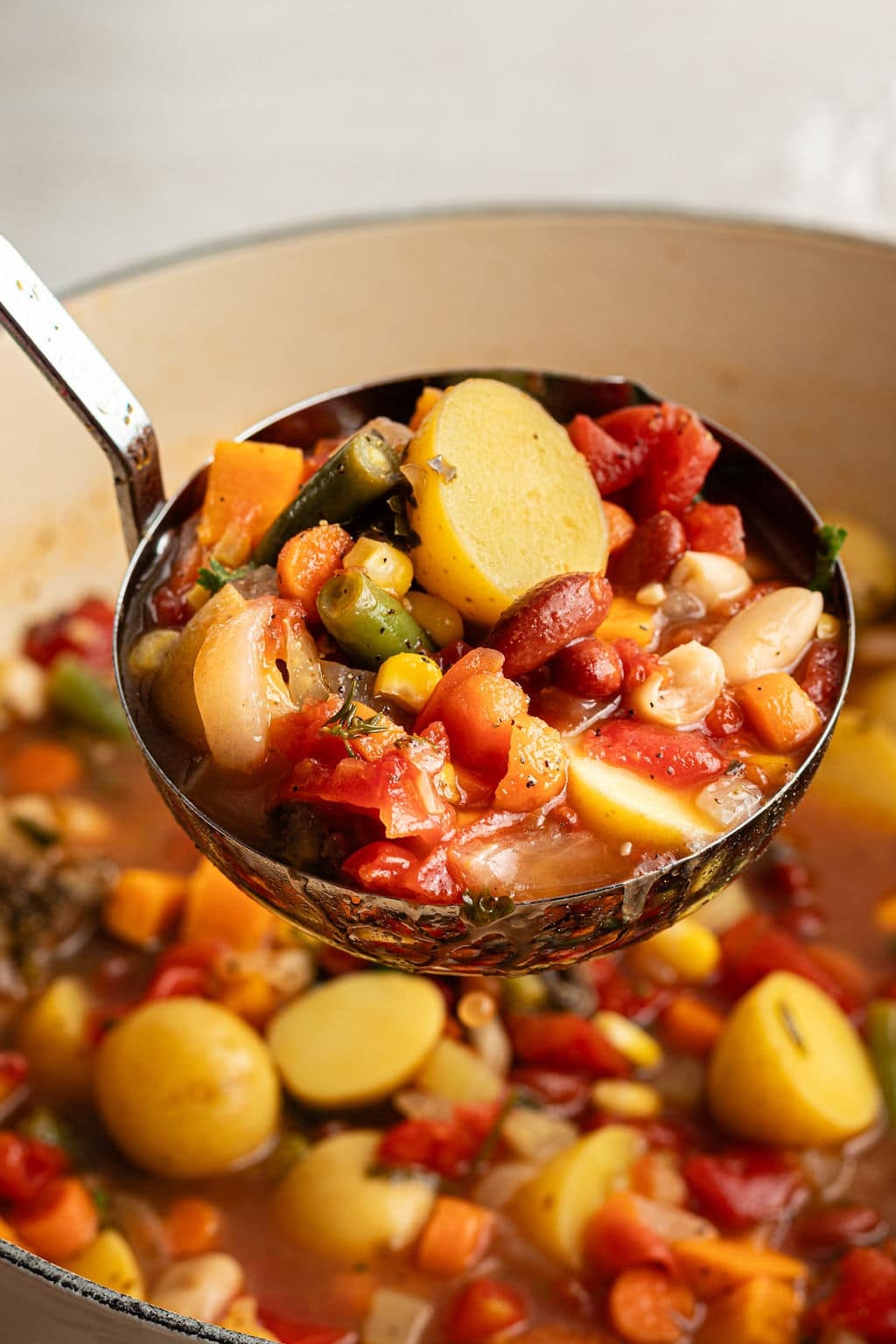 Close-up of a ladle lifting a serving of vegetable soup, showing chunks of potatoes, carrots, green beans, tomatoes, and beans in a rich broth.