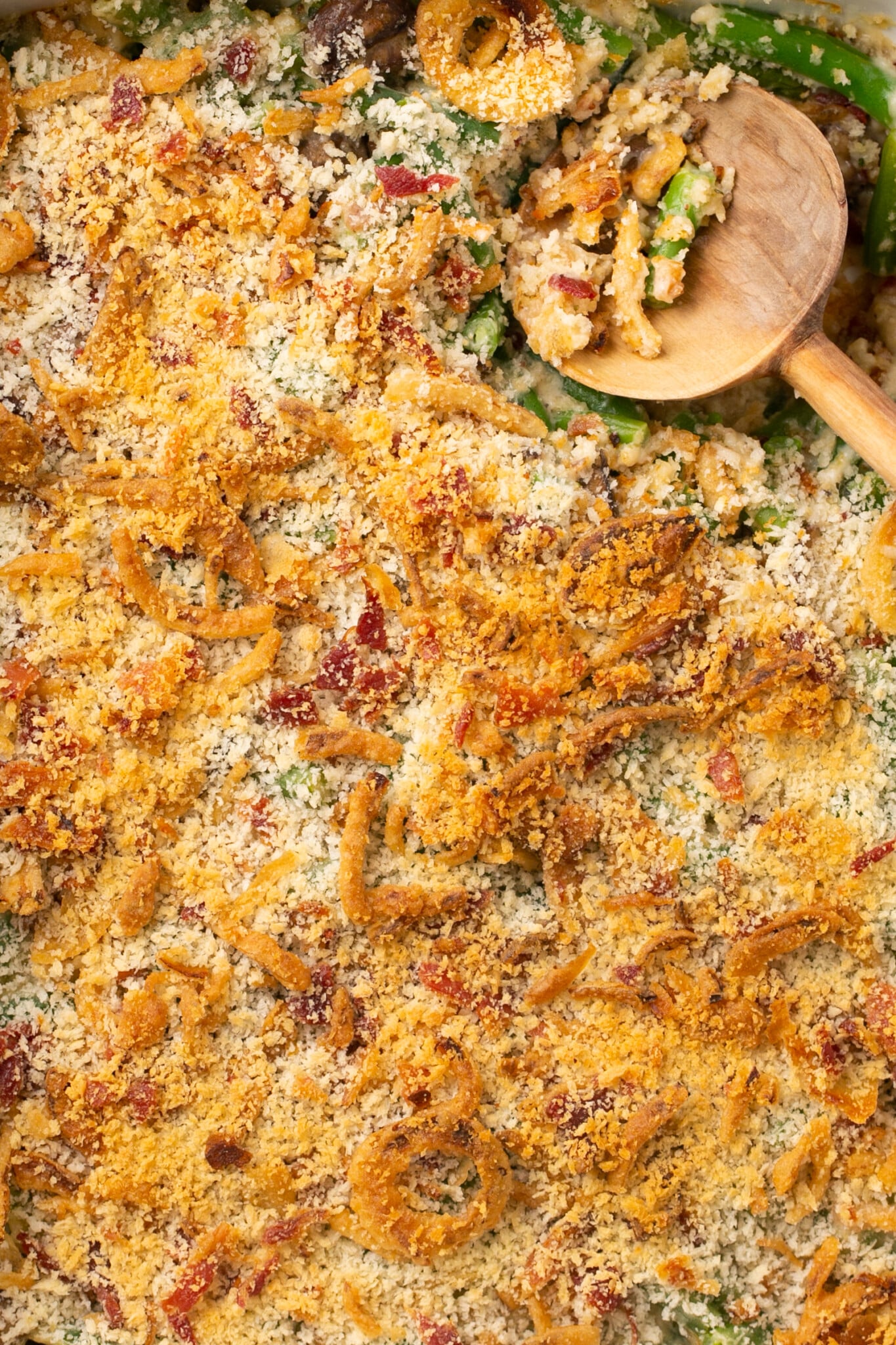 a wooden spoon in a casserole dish made with green beans and topped with panko breadcrumbs and bacon crumbles. 
