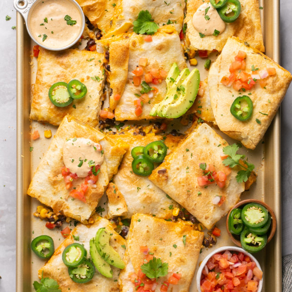 sheet pan with quesadillas cut into squares topped with avocado and jalapeno with two bowls of sauce and salsa