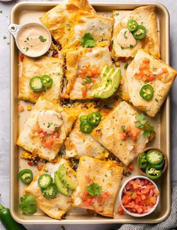 sheet pan with quesadillas cut into squares topped with avocado and jalapeno with two bowls of sauce and salsa