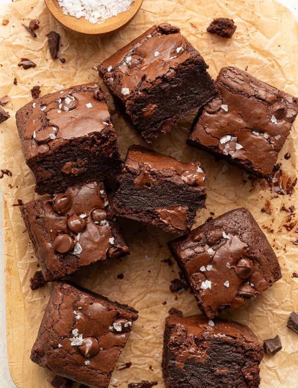 Several fudgy brownie squares, some stacked and one with a bite taken out, are arranged on parchment. The tops are sprinkled with flaky sea salt and melty chocolate chips.