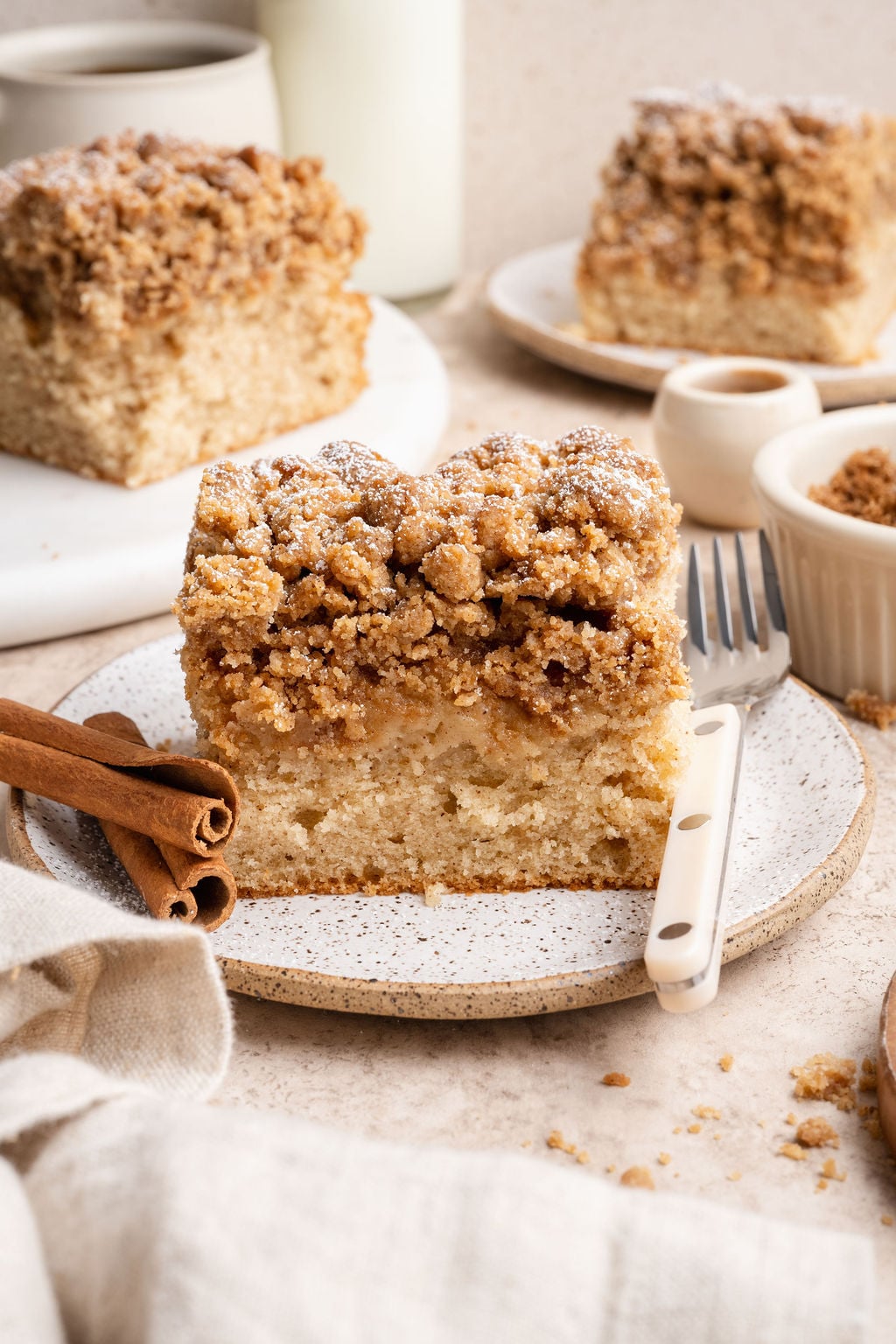 A thick slice of cinnamon crumb cake on a speckled plate, topped with a generous buttery streusel and a light dusting of powdered sugar. Cinnamon sticks and a fork sit beside it, with additional slices of the cake, a cup of coffee, and a small bowl of crumble blurred in the background.
