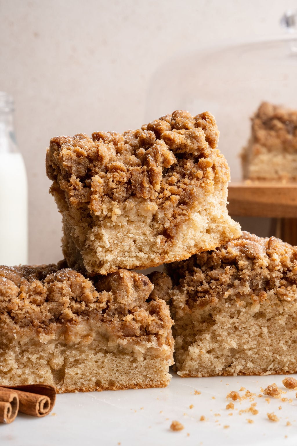 Three pieces of cinnamon crumb cake stacked and slightly offset, showing the fluffy interior and layered crumb topping, with cinnamon sticks and a blurred slice in the background.