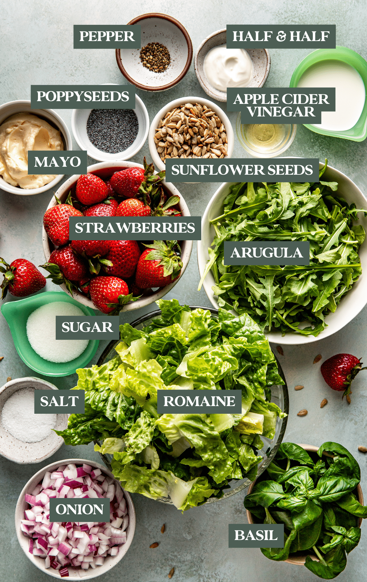 Overhead view of fresh salad ingredients laid out before assembly, including strawberries, arugula, romaine, spinach, red onion, sunflower seeds, poppy seeds, mayo, yogurt, milk, vinegar, and sugar.

