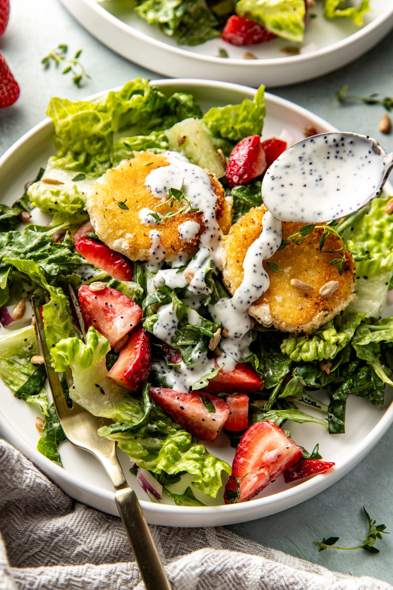 Overhead view of a close-up salad plate with two goat cheese medallions, fresh strawberries, and greens, as a spoon pours creamy poppyseed dressing on top.

