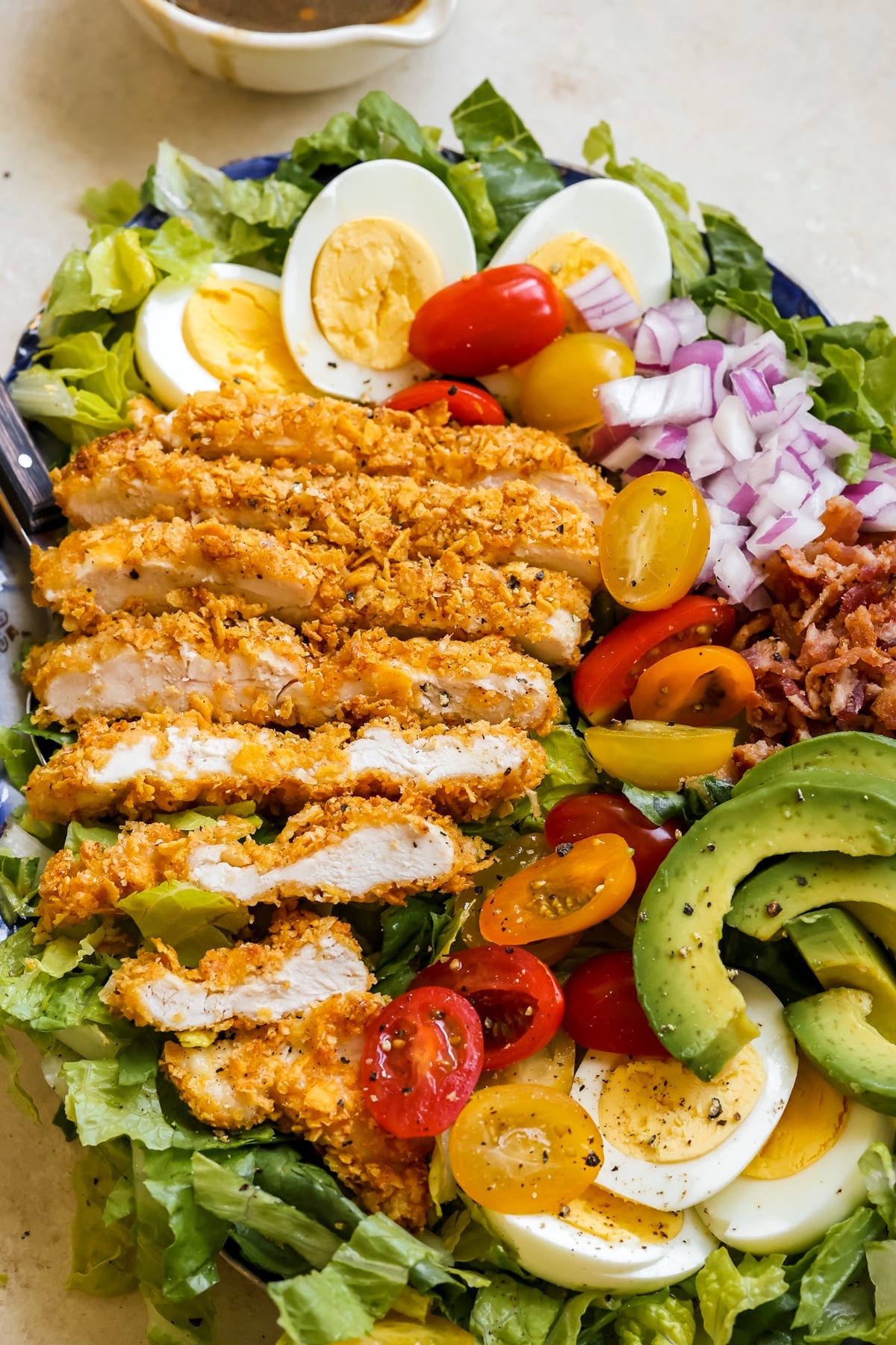romaine lettuce, chicken, avocado, red onion, tomatoes, eggs and bacon on a platter with black pepper on top. 