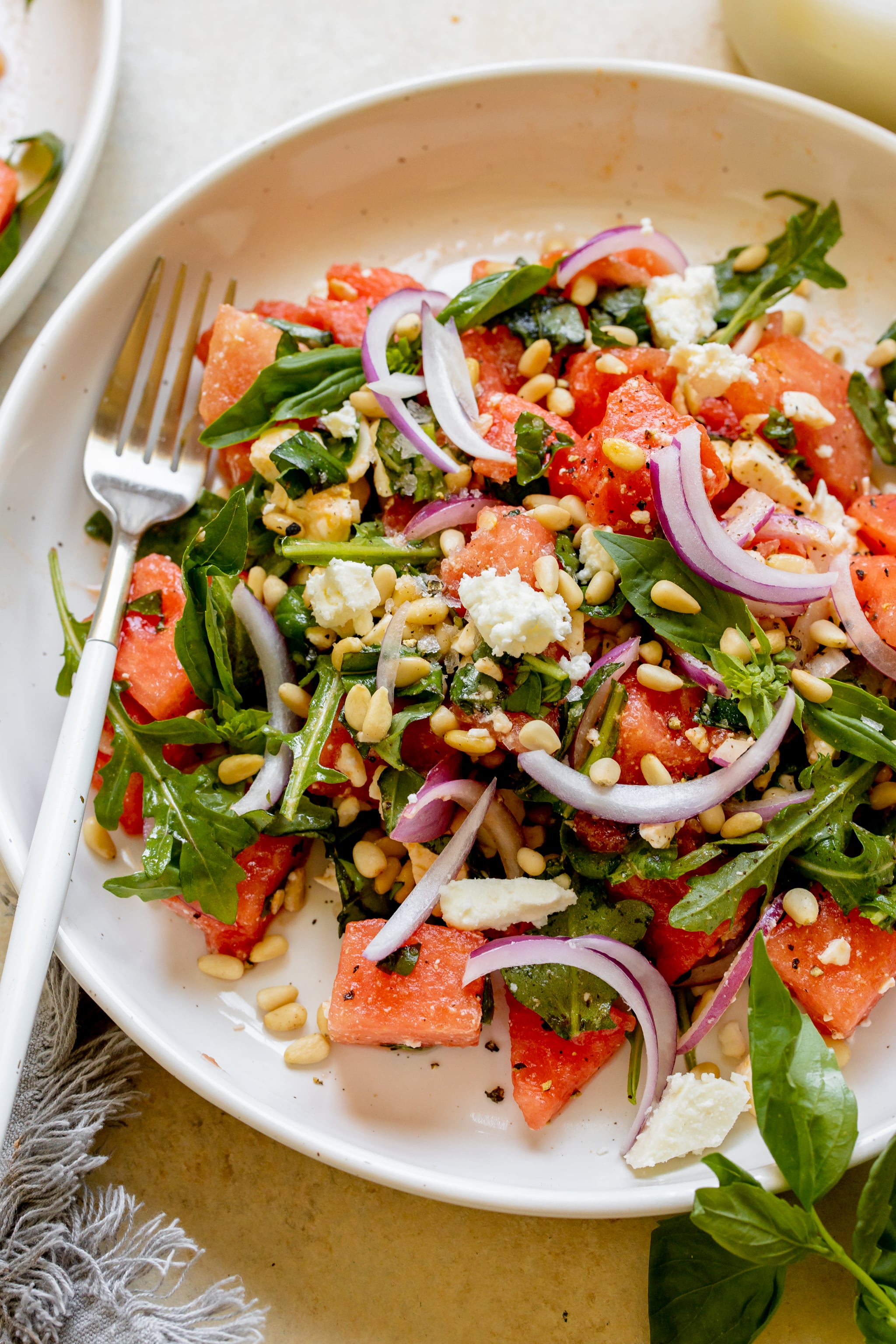 watermelon, arugula, basil, feta, pine nuts and red onion in a white bowl.