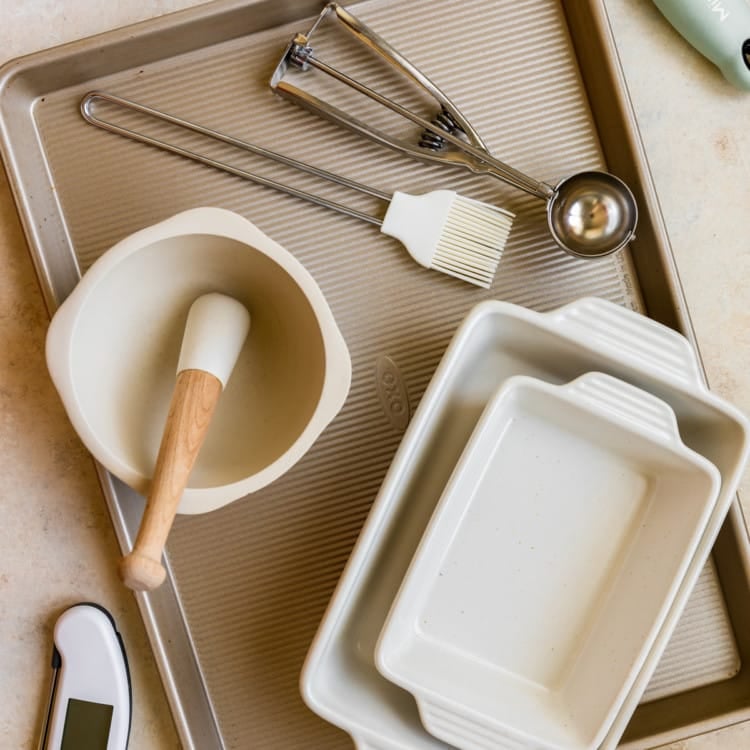gold sheet pan, meat thermometer, yellow juice press, two white casserole dishes, bowl, brush, and cookie scoop