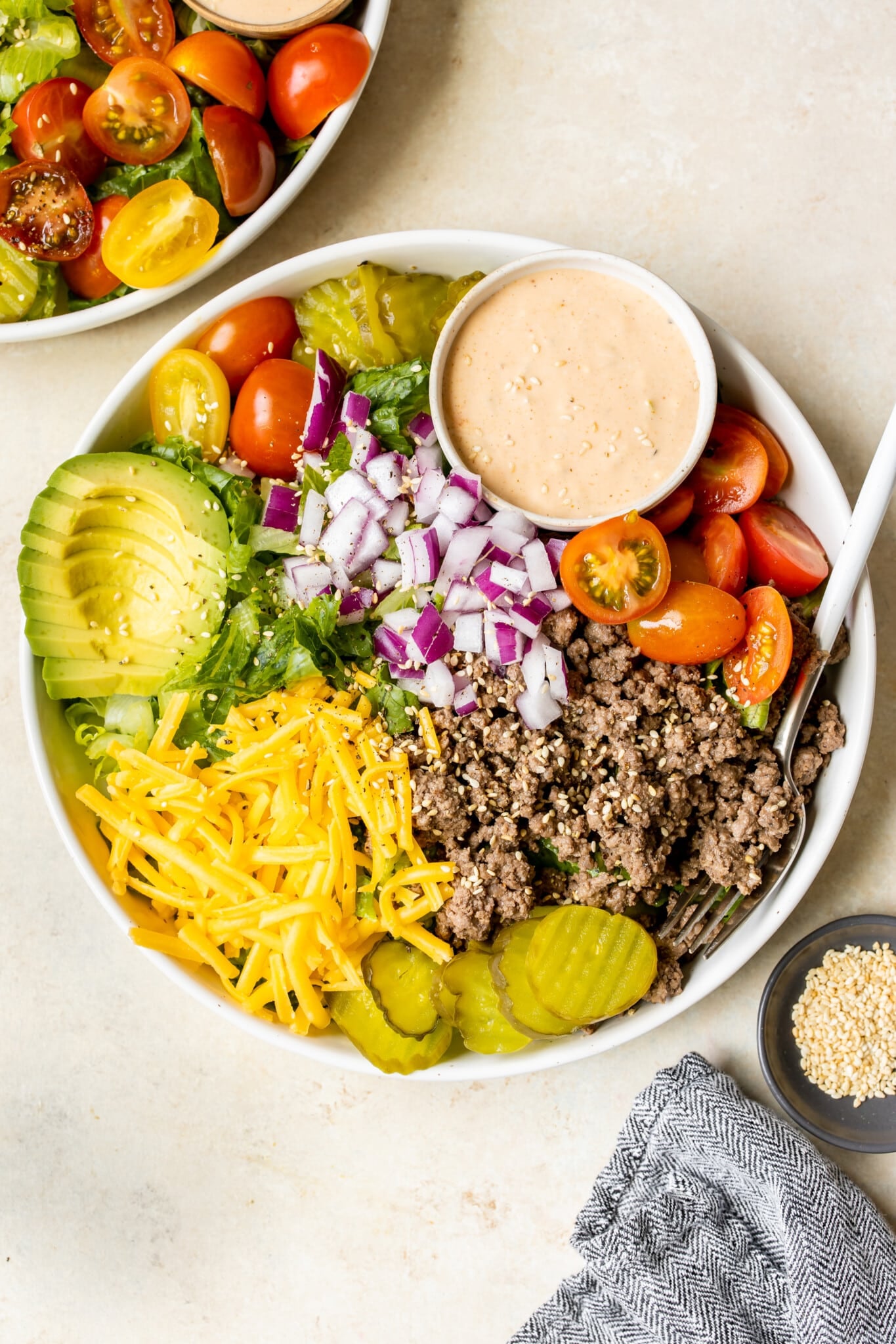 romaine lettuce in a white bowl topped with burger toppings like ground beef, cheese, avocado, red onion, pickles, tomatoes and a special burger sauce on the side. 
