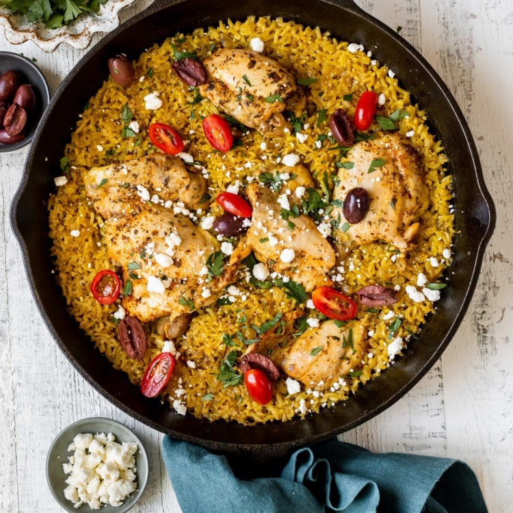 black cast iron skillet with yellow rice, chicken thighs, cherry tomatoes, feta cheese, kalamata olives, and green spices
