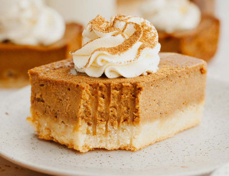 one slice of pumpkin pie bars with a bite missing on a tan plate and topped with whipped cream and a dust of spices