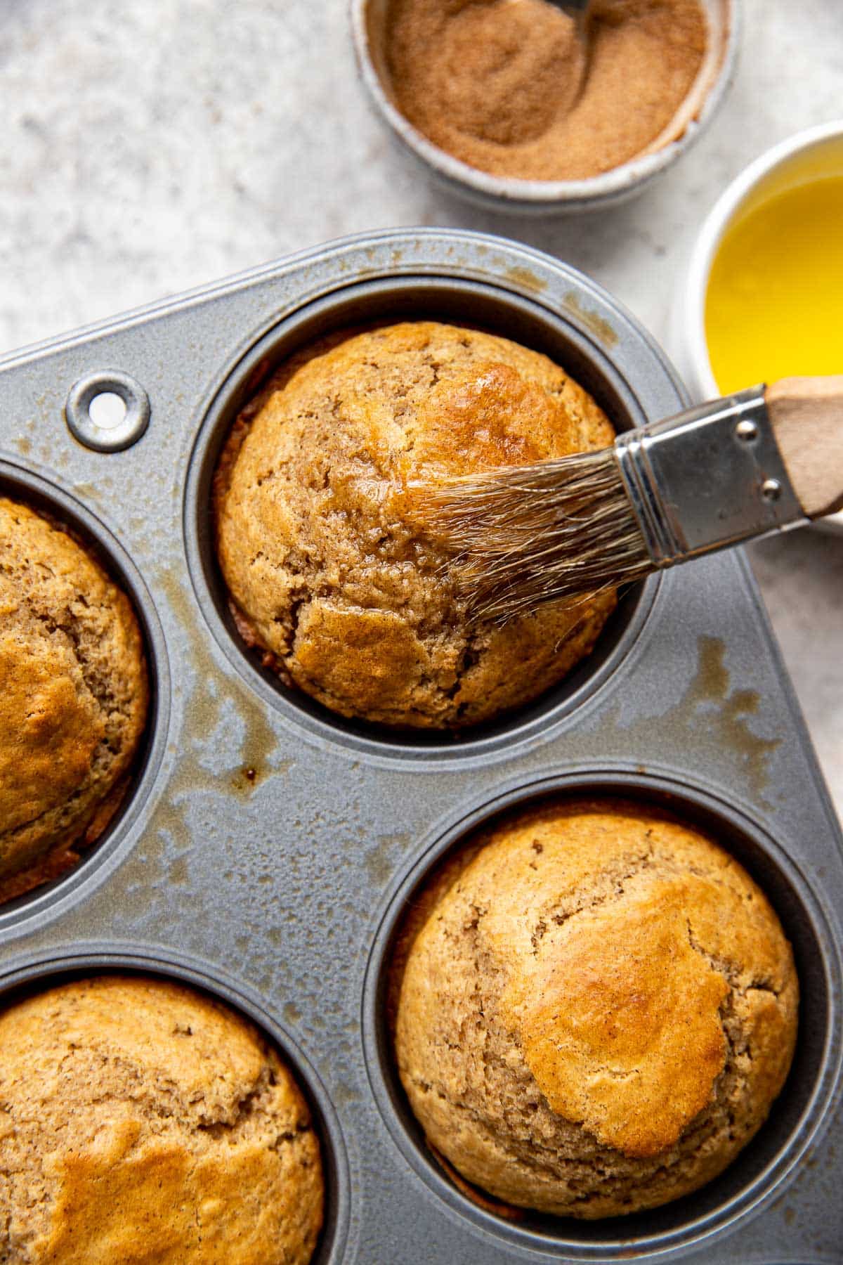 butter being brushed on top of muffins in a muffin pan. 