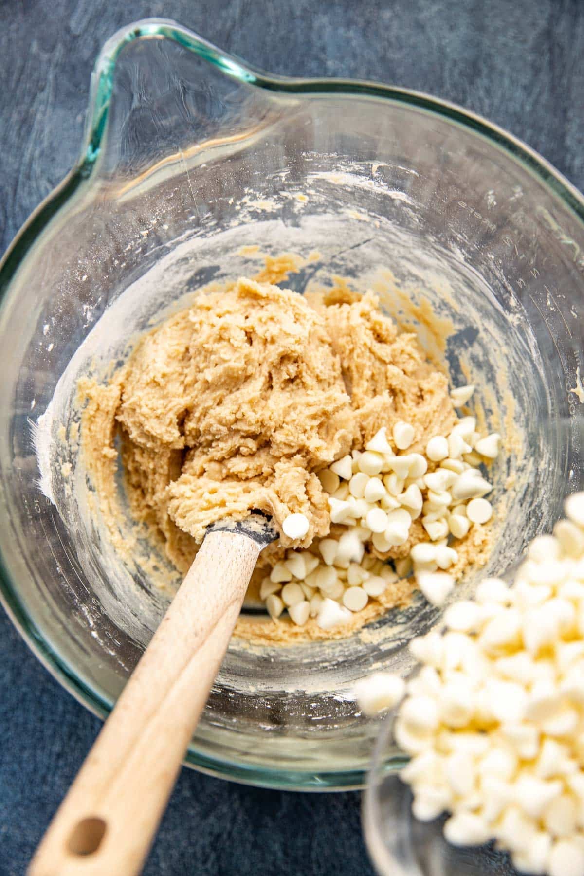 white chocolate chips being poured into sugar cookie dough in a glass stand mixing bowl. 