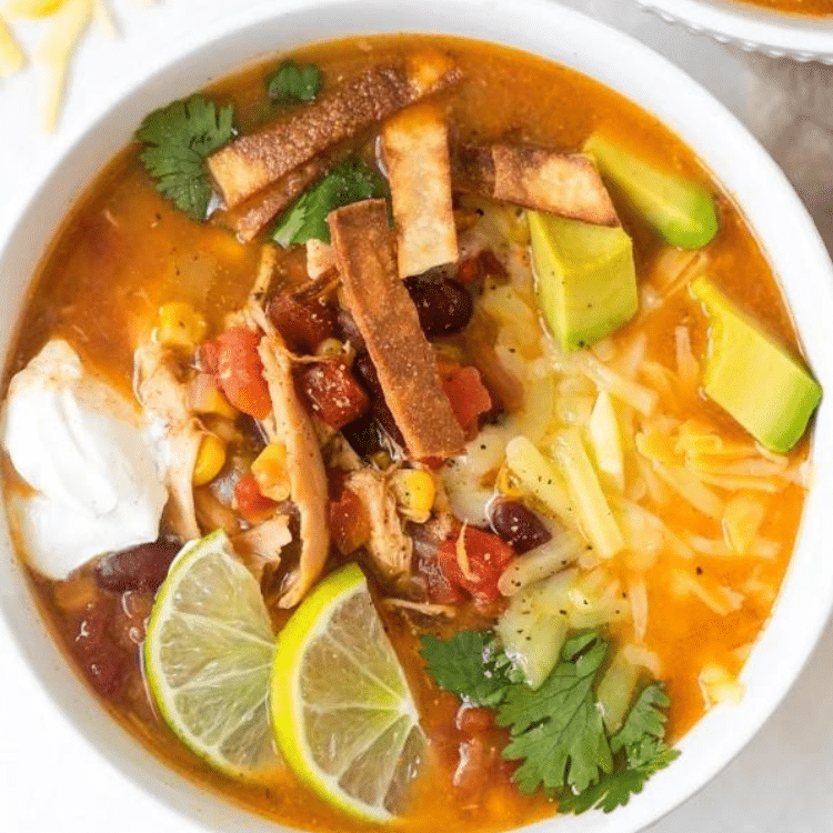 white bowl of taco soup with sour cream, avocado, cheese, tortilla strips, and limes