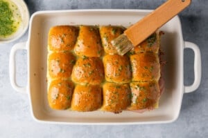 melted butter and parsley being brushed on top of slider buns.