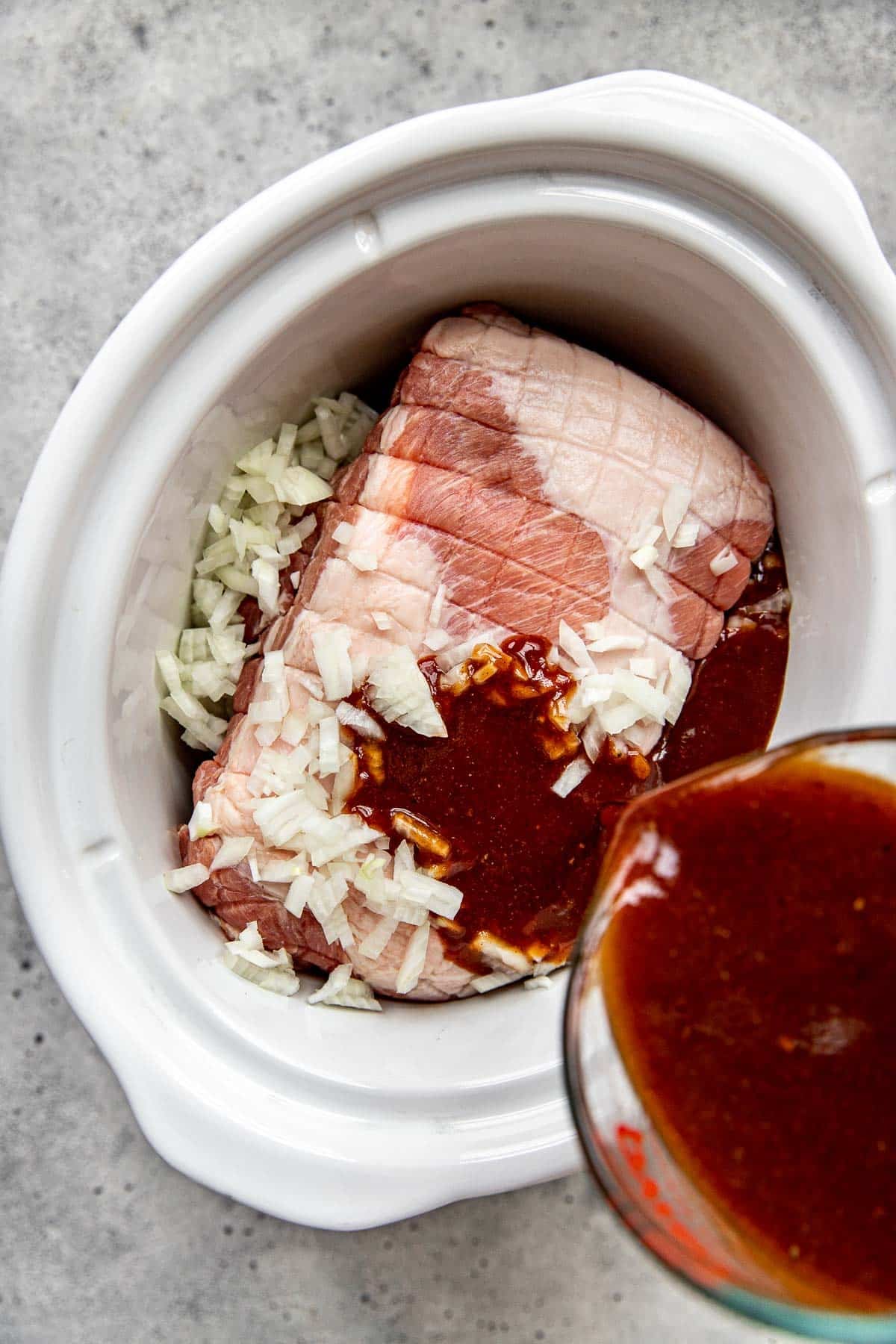 Sauce being poured onto pulled pork in a white slow cooker. 
