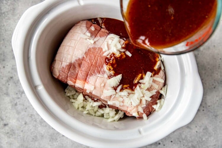 sauce being poured on a boneless pork butt in a white slow cooker.