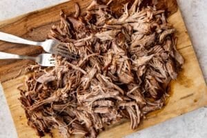 two forks shredding pulled pork on a wooden cutting board.