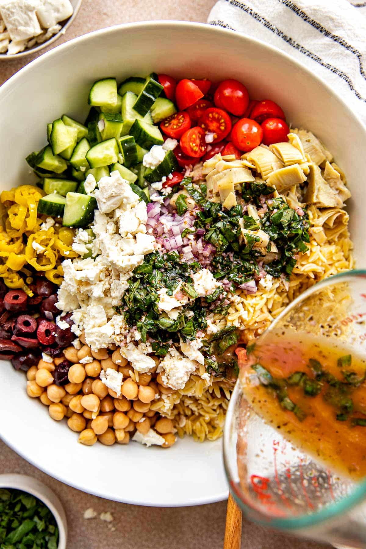 orzo salad in a large white bowl with dressing being poured on top. 