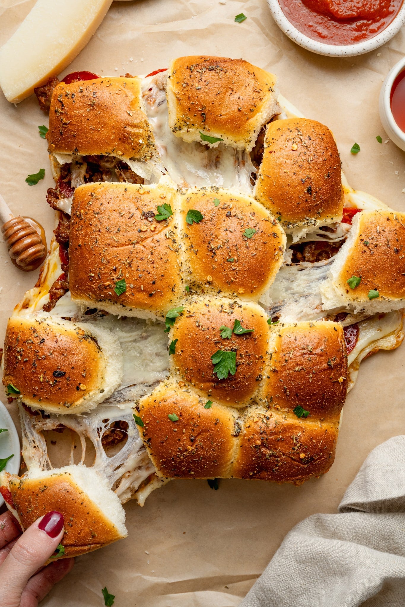 Overhead view of freshly baked pizza sliders being pulled apart, showing gooey melted cheese stretching between the rolls. The golden tops are brushed with garlic butter and sprinkled with Italian herbs and parsley, with bowls of marinara sauce nearby for dipping.