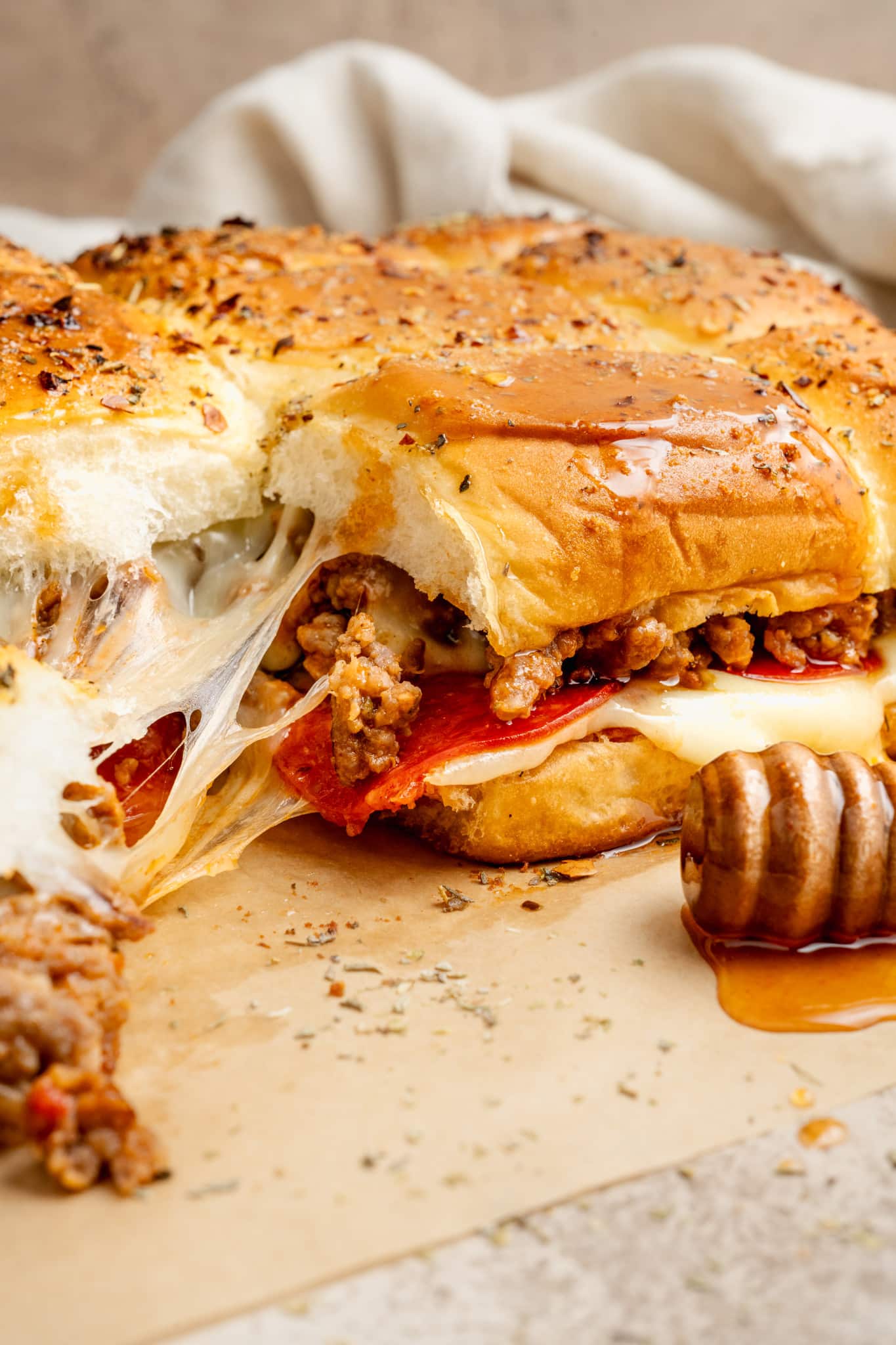 Side view of pizza sliders filled with melted mozzarella, pepperoni, and sausage. The tops are brushed with honey and garlic butter, and a honey dipper rests nearby on the parchment paper.