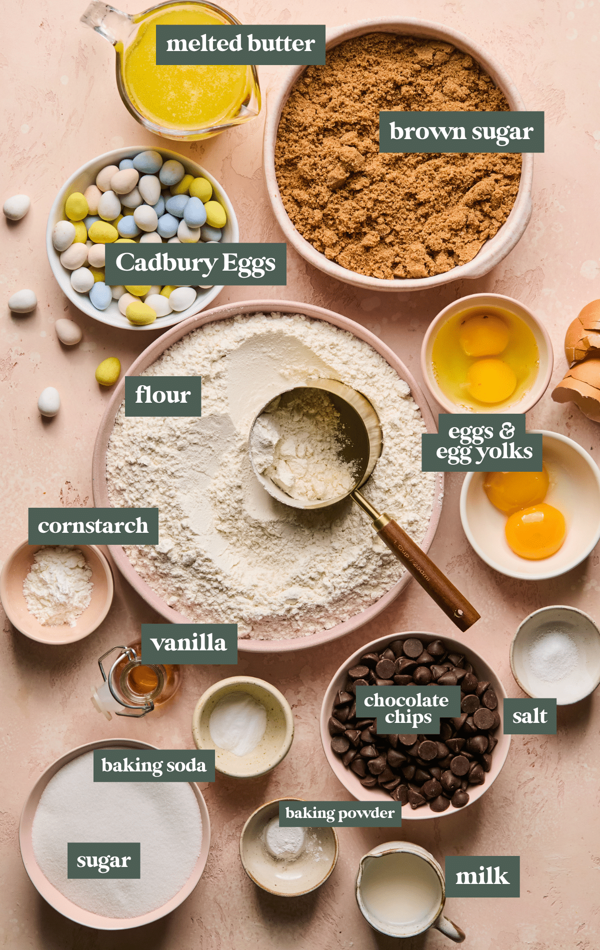 Flat lay of labeled ingredients for Easter sheet pan cookies, including melted butter, brown sugar, Cadbury Mini Eggs, flour, eggs and egg yolks, cornstarch, vanilla, sugar, baking soda, baking powder, salt, chocolate chips, and milk—all arranged in bowls on a blush pink background.