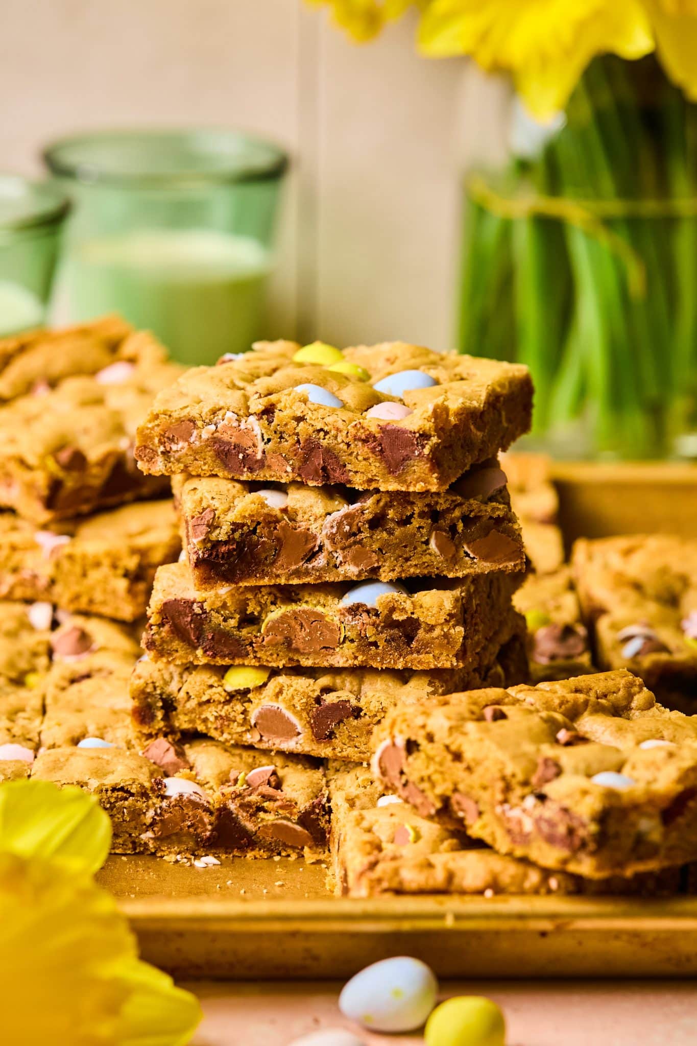 Stack of thick, chewy Easter cookie bars with chocolate and pastel candy eggs, in front of blurred spring-themed background.