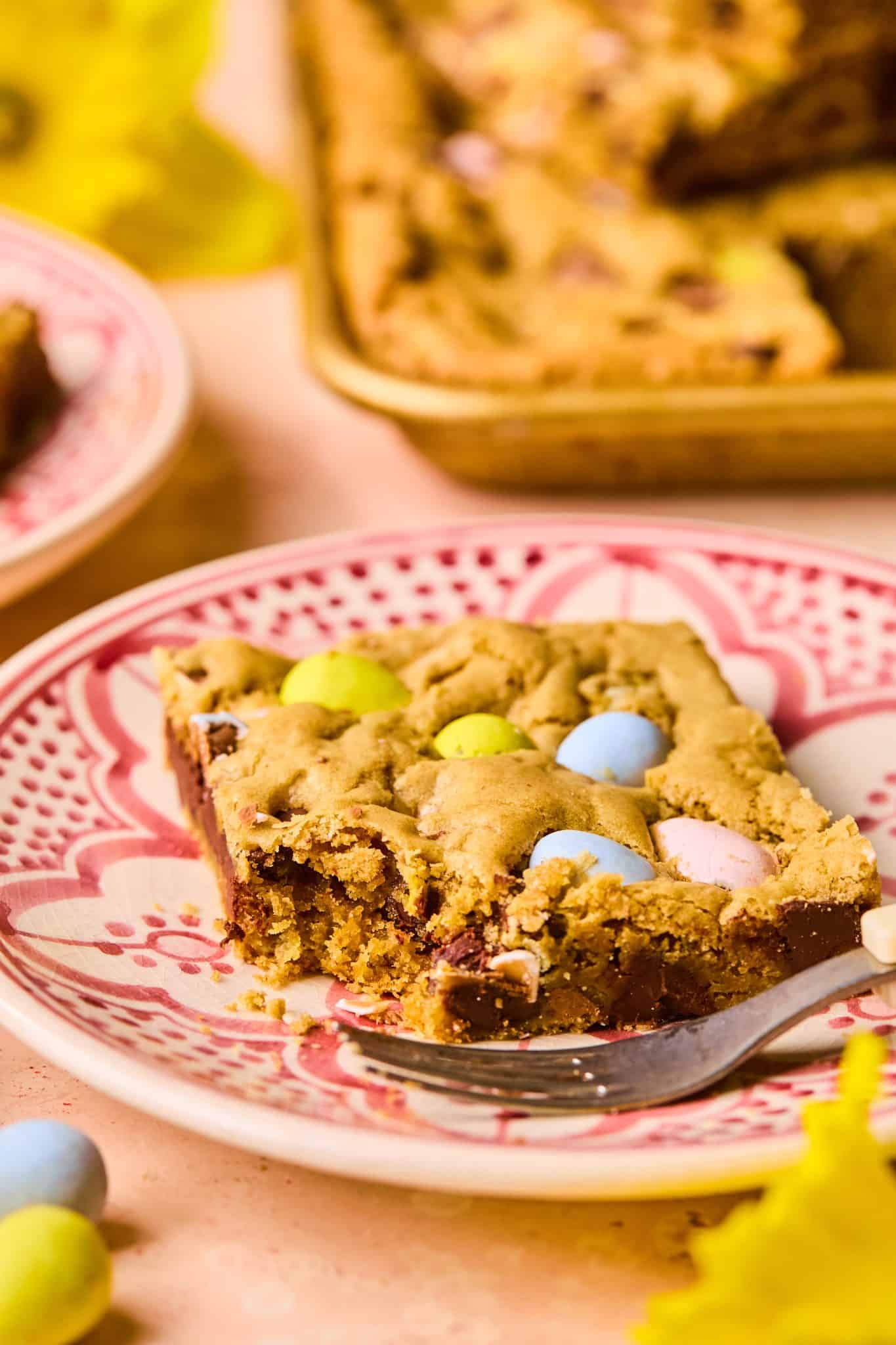 A Easter cookie bar on a pink patterned plate with a bite taken out, surrounded by festive spring decor.