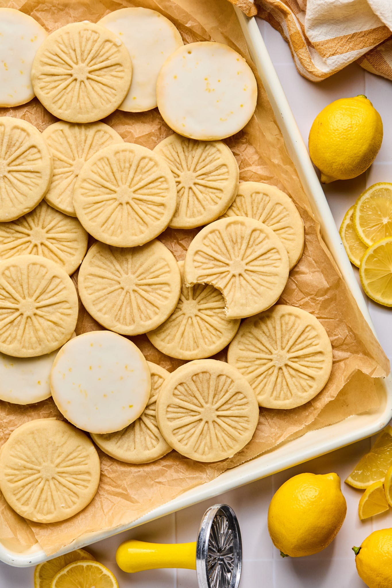 Picture of lemonade cookies on parchment-lined baking sheet. One cookie has a bite taken out of it. There are lemons and the metal lemon stamp surrounding the baking sheet.