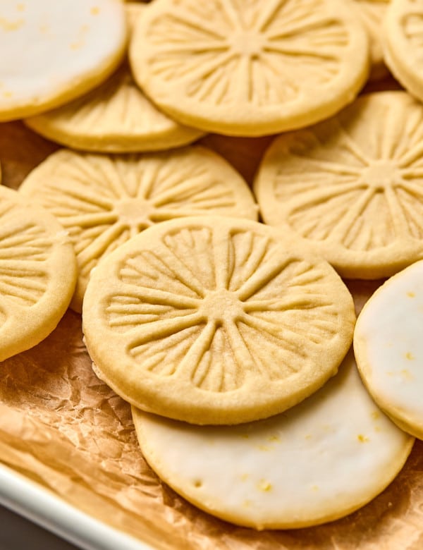 Up close picture of lemon cookies on parchment-lined baking tray. Picture shows both sides of the cookies as some are turned upright with the lemon stamp and some are flipped over to show the lemon glaze on the bottom.