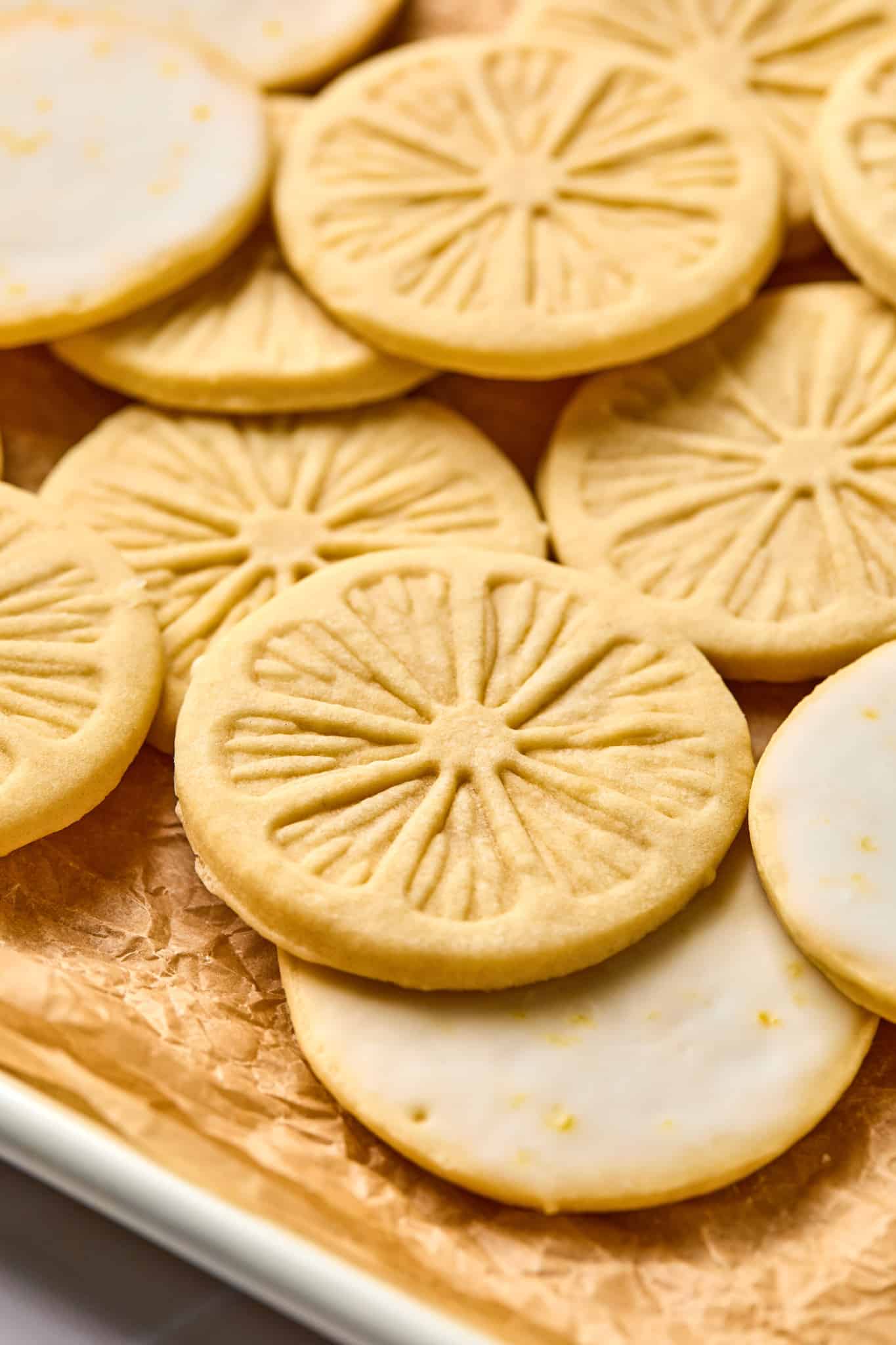 Up close picture of lemon cookies on parchment-lined baking tray. Picture shows both sides of the cookies as some are turned upright with the lemon stamp and some are flipped over to show the lemon glaze on the bottom.