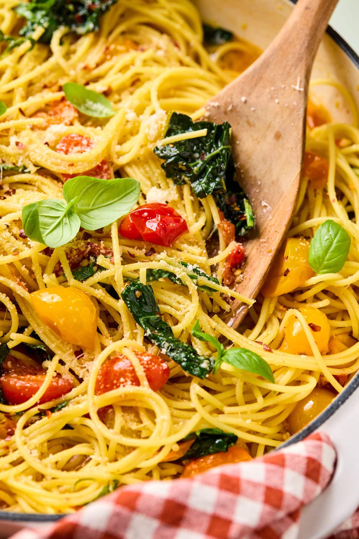  A white cast-iron skillet filled with spaghetti, cherry tomatoes, and greens, being stirred with a wooden spoon, with fresh basil leaves on top.
