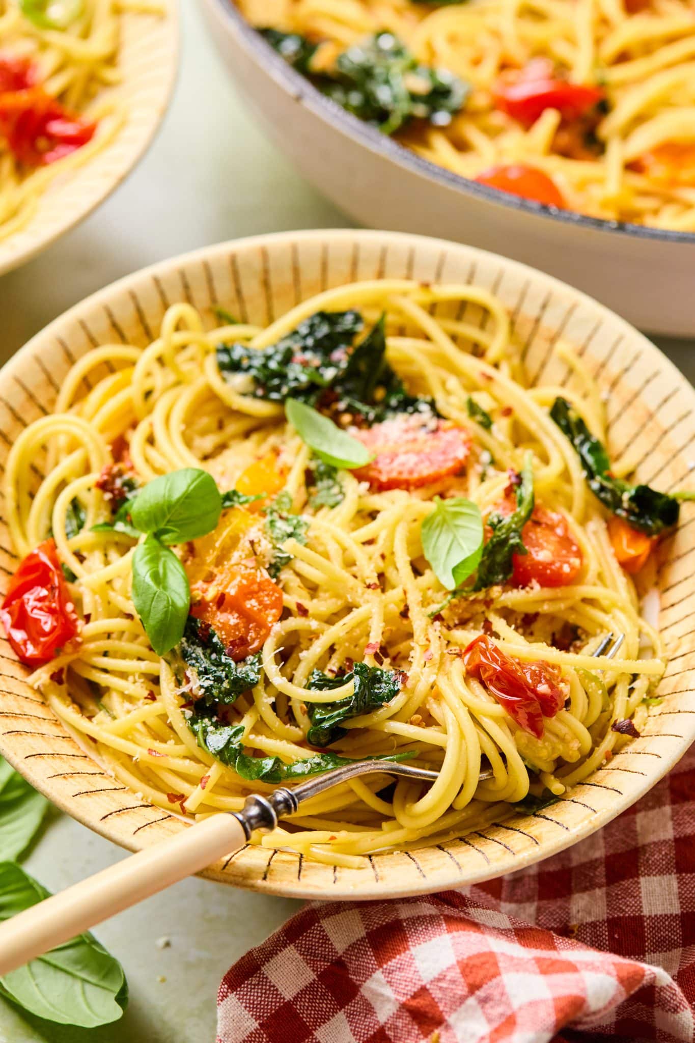 Two bowls of spaghetti with sautéed cherry tomatoes and dark green kale, garnished with fresh basil, placed on a table with a red-and-white checkered napkin.