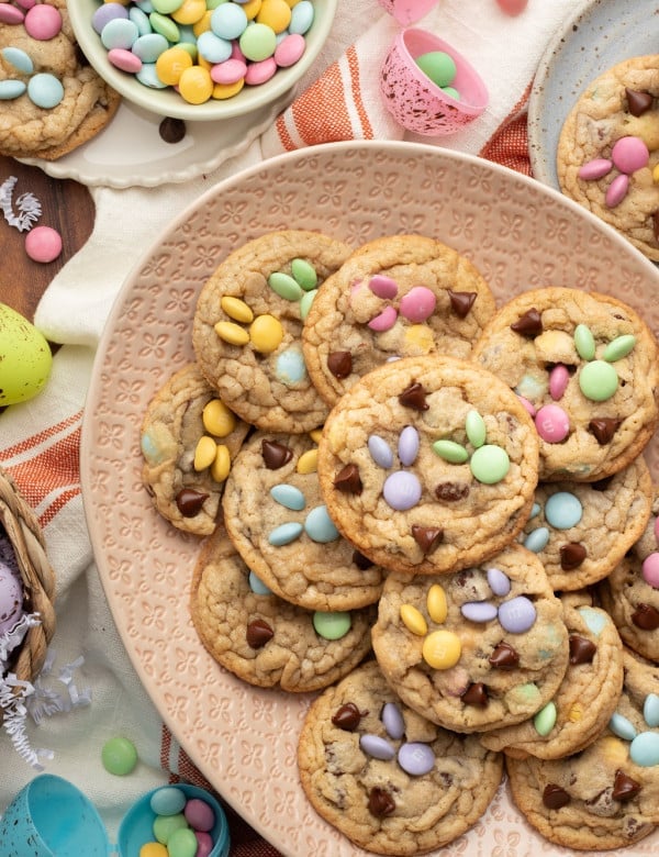 A decorative pink plate filled with spring-themed M&M cookies, featuring pastel-colored candies arranged in bunny designs. A bowl of extra M&Ms and festive Easter decorations surround the plate.