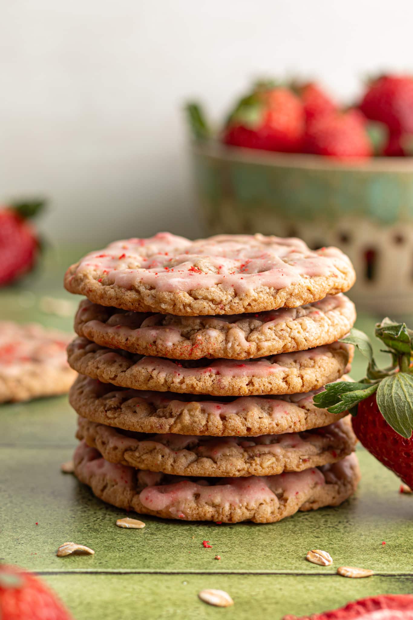 A stack of six strawberry-glazed oatmeal cookies on a green surface, with fresh strawberries and oats scattered around. A bowl of strawberries is softly blurred in the background.
