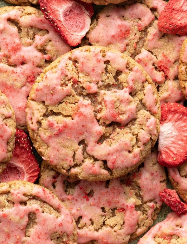 A close-up overhead shot of strawberry-glazed oatmeal cookies, showing their craggy texture and pink glaze. Freeze-dried strawberry slices are scattered among the cookies.