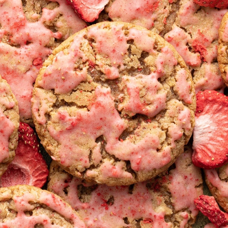 A close-up overhead shot of strawberry-glazed oatmeal cookies, showing their craggy texture and pink glaze. Freeze-dried strawberry slices are scattered among the cookies.