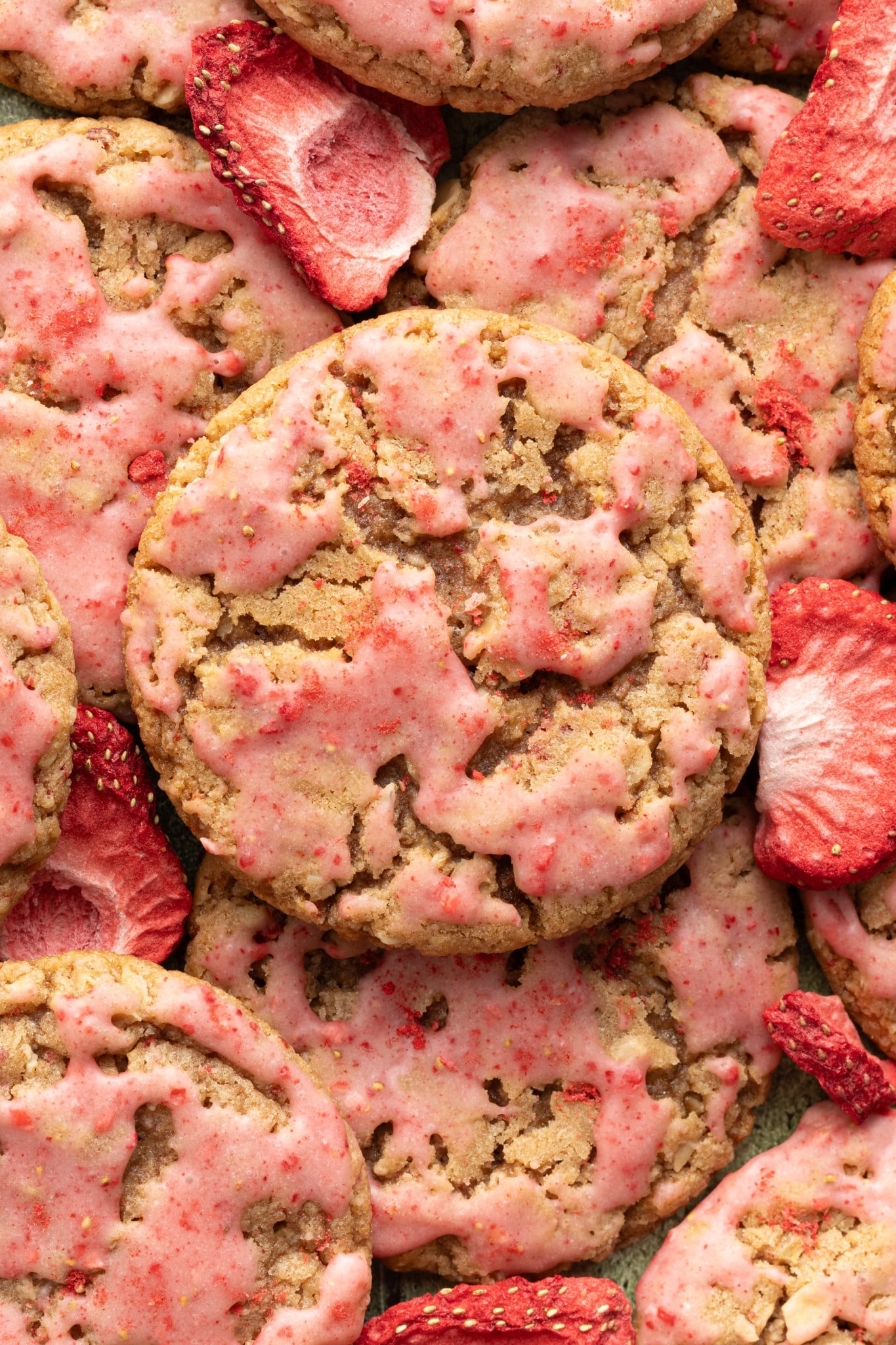 A close-up overhead shot of strawberry-glazed oatmeal cookies, showing their craggy texture and pink glaze. Freeze-dried strawberry slices are scattered among the cookies.
