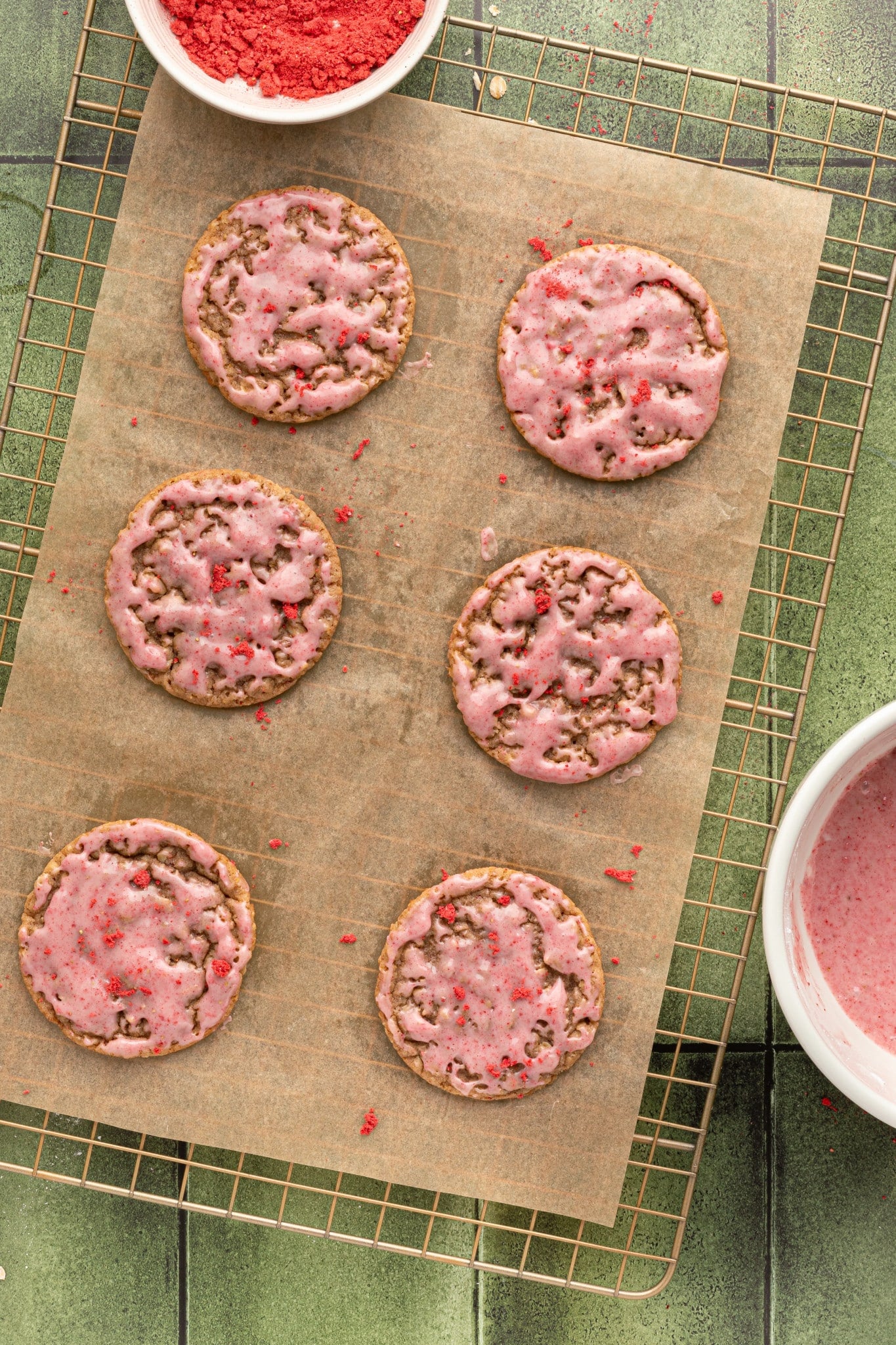 Six strawberry-glazed oatmeal cookies on a parchment-lined wire rack, viewed from above. A bowl of strawberry powder and some glaze are visible in the corners.