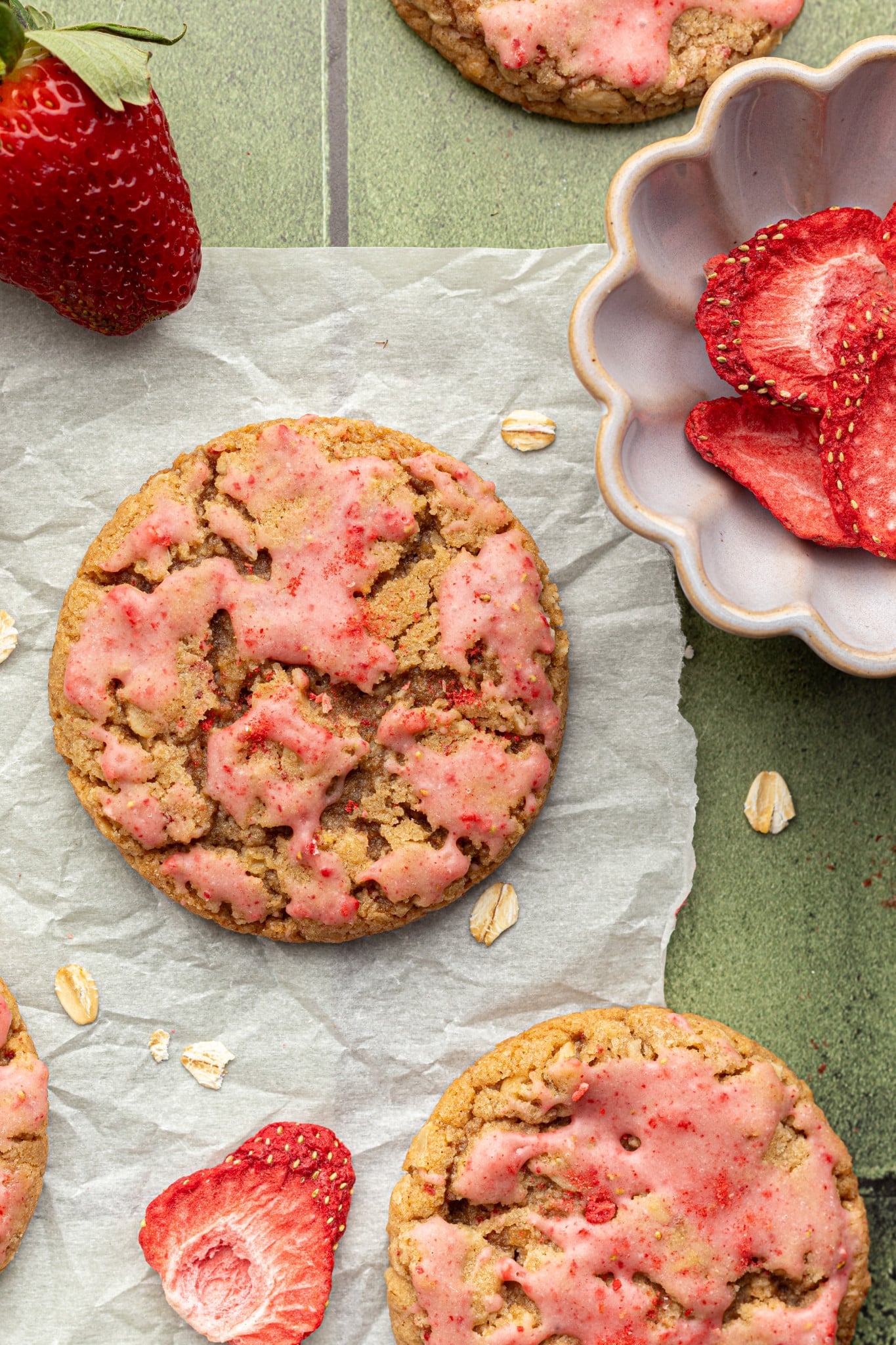 Top-down view of strawberry oatmeal cookies on parchment paper, with whole and sliced strawberries and oats around them. A fluted bowl holds freeze-dried strawberry slices.