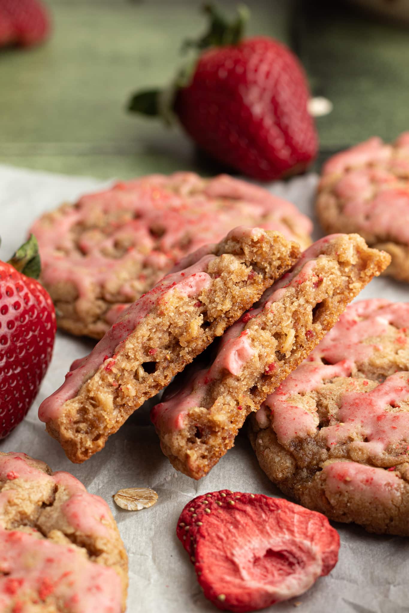 A halved cookie showing the chewy interior of a strawberry-glazed oatmeal cookie. Surrounded by whole and freeze-dried strawberries on parchment.